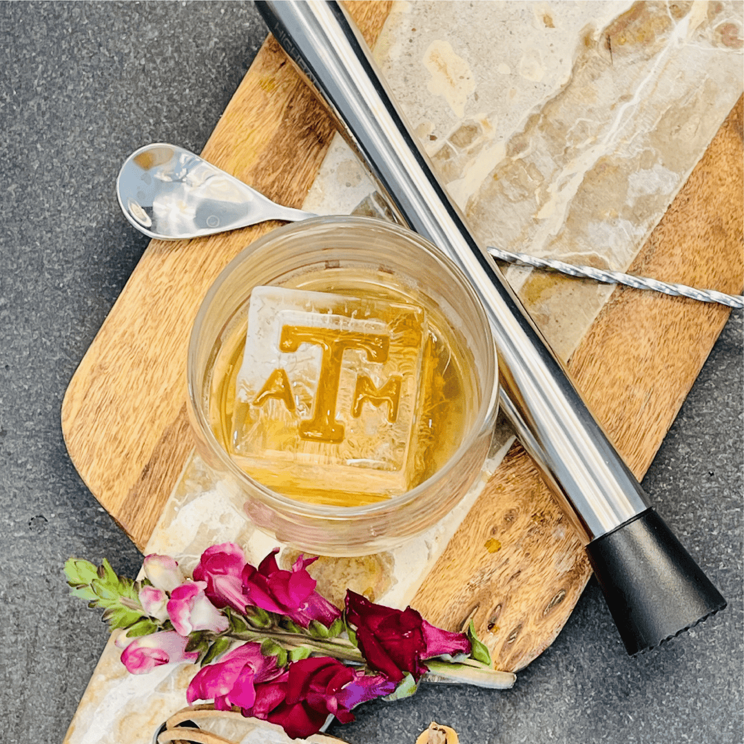 A glass of amber liquid with a large, clear ice cube featuring Texas A&M Block ATM Inserts - 3 Pack embossed with the letters ATM.