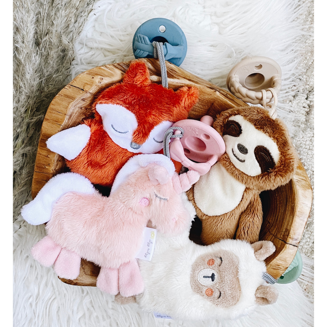 A variety of colorful baby toys including Sweetie Pal™ Plush & Pacifier: Dino pacifiers and teething rings neatly arranged in a wooden bowl on a plush white rug, featuring animal designs like a sloth and a fox.