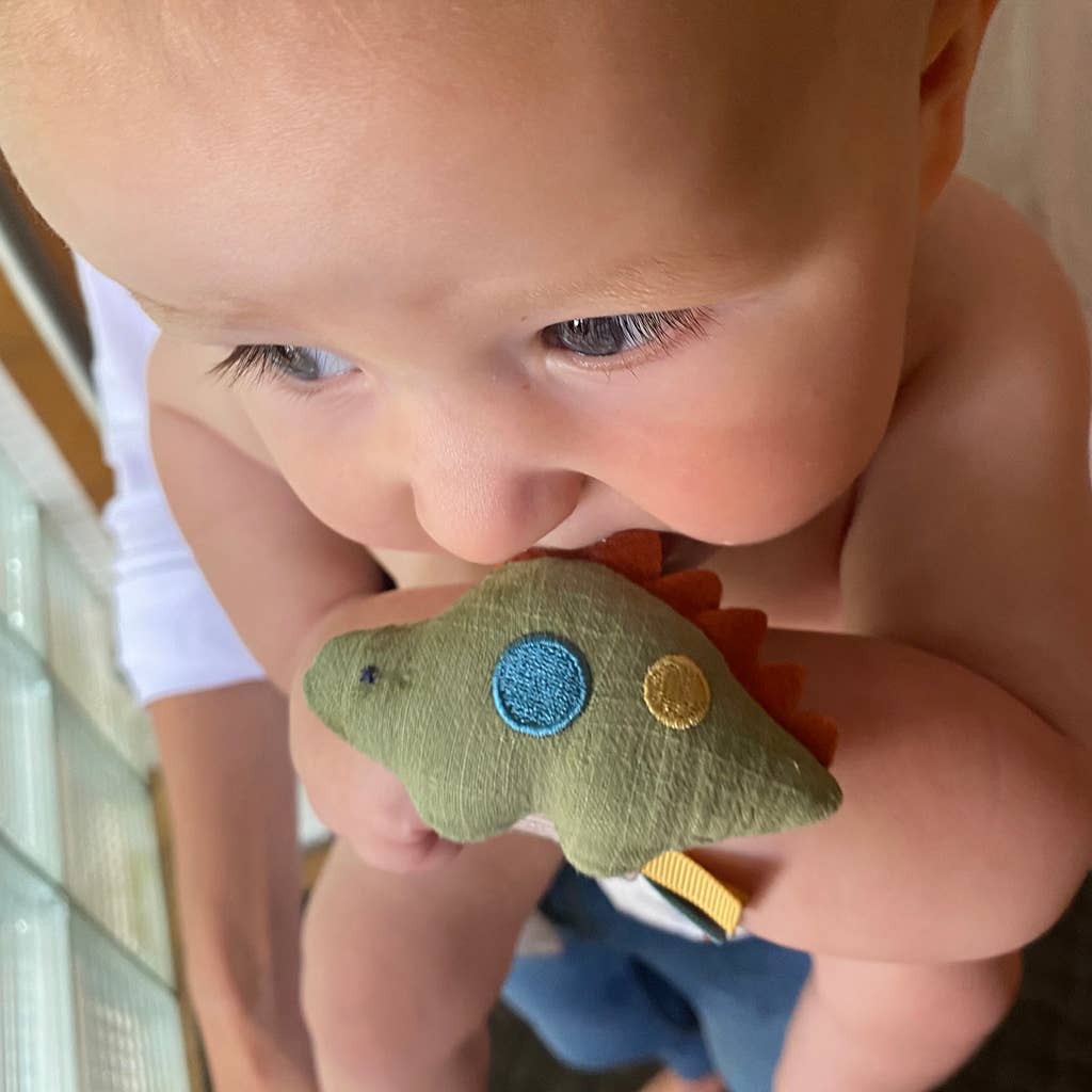 A close-up of a baby holding a small Itzy Bitzy Wrist Rattle: Cottage toy in its natural cotton fabric hands, examining it with a curious expression. The toy is green with colorful patches.