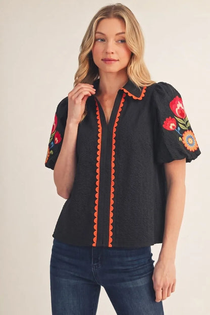 Fall in Bloom Blouse: black top with colorful floral embroidery on sleeves and orange scalloped trim. Available in sizes XS-XL.