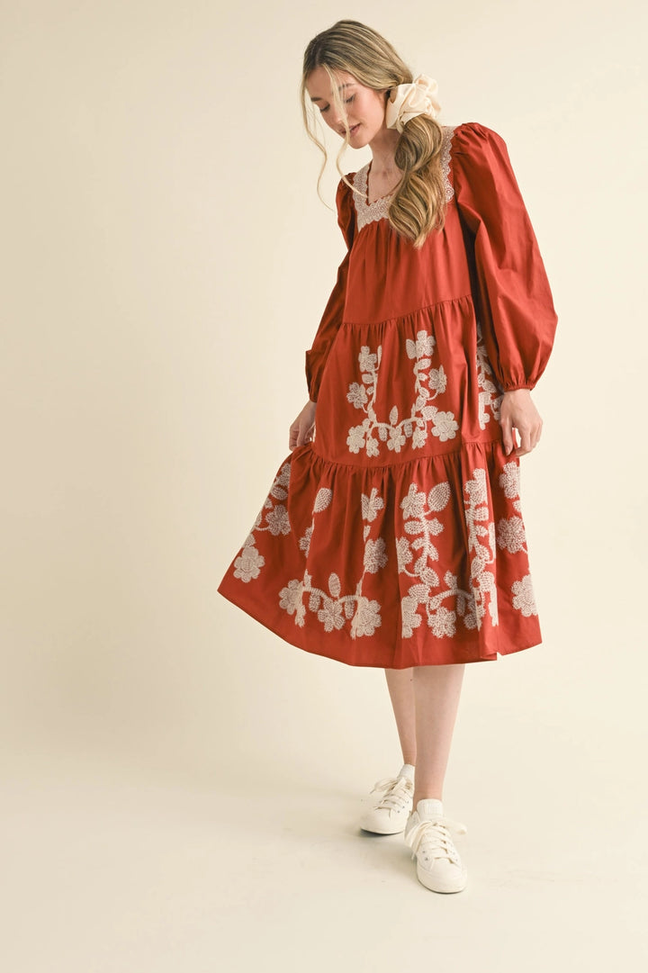 Rust tiered midi dress with square neckline, long puff sleeves, and white floral embroidery. Available in all sizes.
