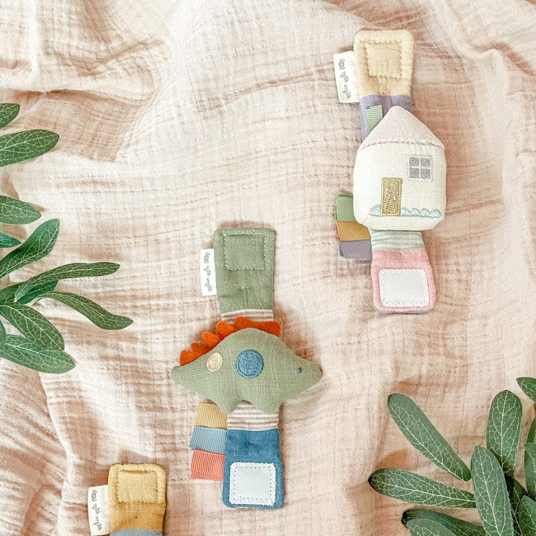 Itzy Bitzy Wrist Rattle: Cottage houses and trees made from natural cotton fabric are laid out on a textured beige cloth, surrounded by green plant leaves. The items feature pastel colors and playful designs.