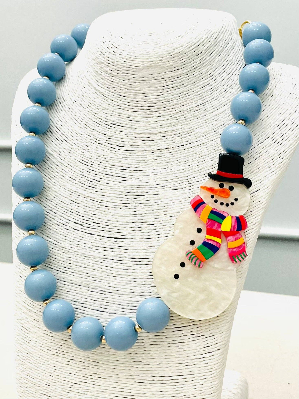 HOLIDAY - Beaded Snowman Necklace (blue/white) BNK1216 features large light blue beads and a snowman pendant with a black hat and colorful striped scarf. Sizes available: not specified.