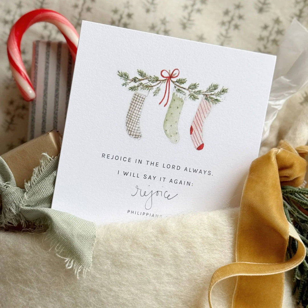 Christmas card with three stockings, greenery, candy cane, and gold ribbon. Reads “Rejoice in the Lord always.” Sizes available.