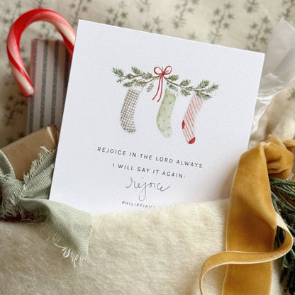 Christmas card with three stockings, greenery, candy cane, and gold ribbon. Reads “Rejoice in the Lord always.” Sizes available.