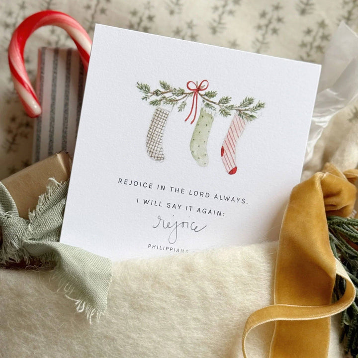 Christmas card with three stockings, greenery, candy cane, and gold ribbon. Reads “Rejoice in the Lord always.” Sizes available.