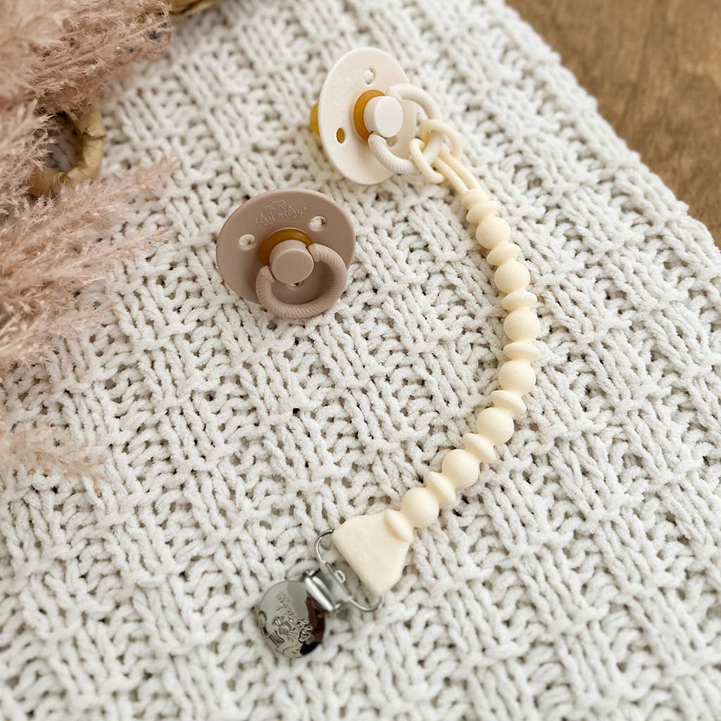 Two Sweetie Strap™ Silicone One-Piece Pacifier Clips with food-grade silicone and wooden beads clip attachment on a textured knit blanket. One pacifier is beige and the other is grey, both with neutral tones.