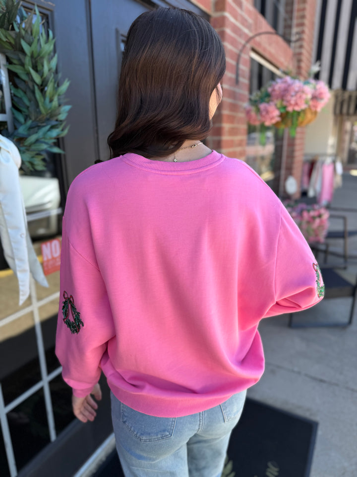 Bright pink Sarah Joy Wreath sweatshirt with embroidered Christmas wreath sleeve details. Available in all sizes.