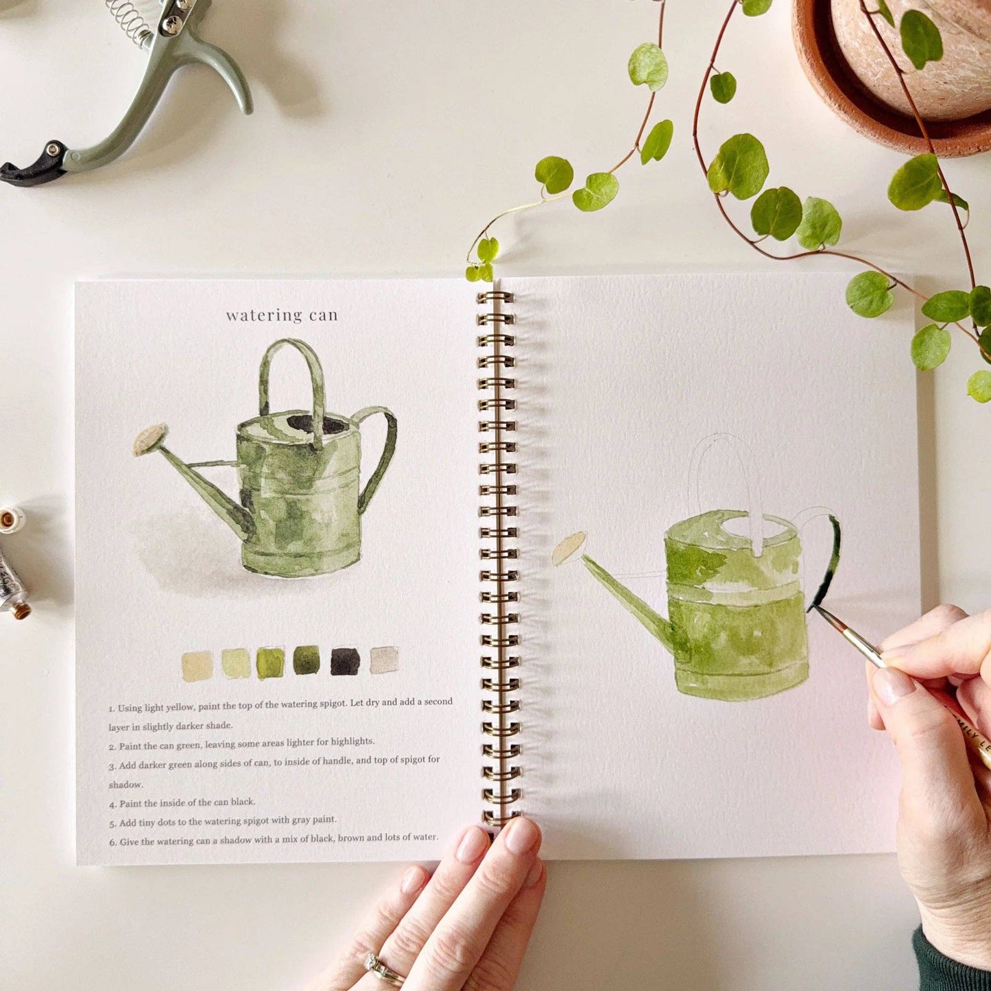 Garden Watercolor Workbook in hand, open to a garden tutorial page. Available sizes: standard and compact.