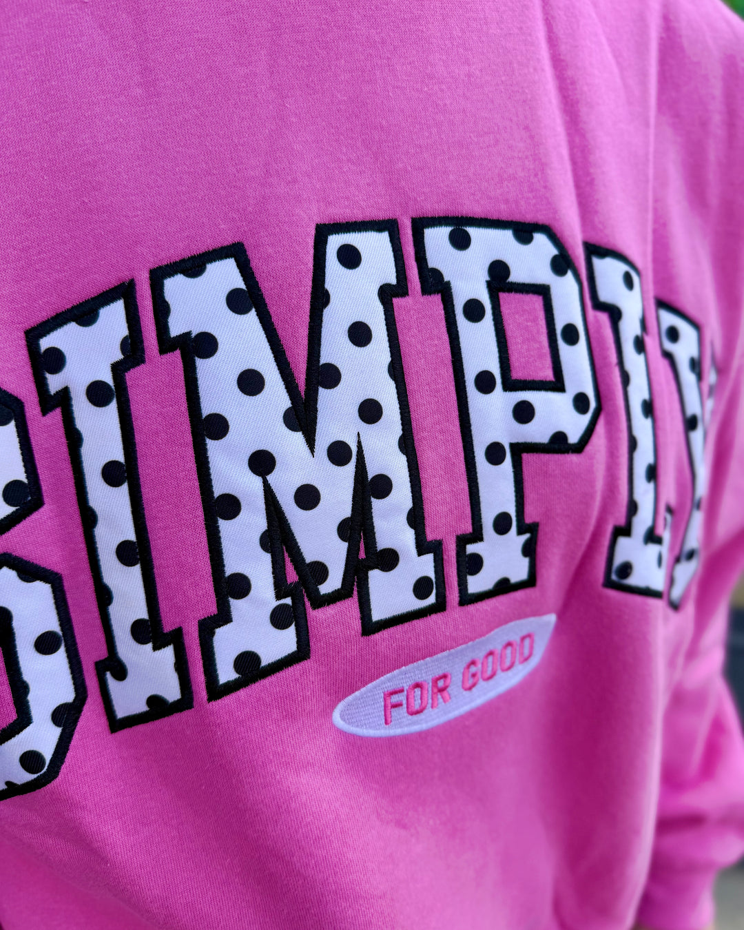 Pink cropped crew neck shirt with large polka-dotted varsity lettering spelling “SIMPLY” and smaller text below reading “FOR GOOD.” Available in sizes XS–XL.