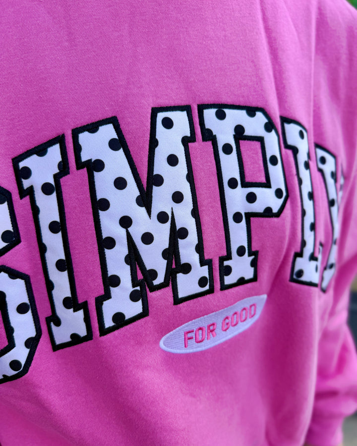 Pink cropped crew neck shirt with large polka-dotted varsity lettering spelling “SIMPLY” and smaller text below reading “FOR GOOD.” Available in sizes XS–XL.