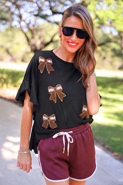 Sideline Sparkle Ruffle Top: black shirt with football bow patch accents and ruffle details. Available in sizes XS–XL.