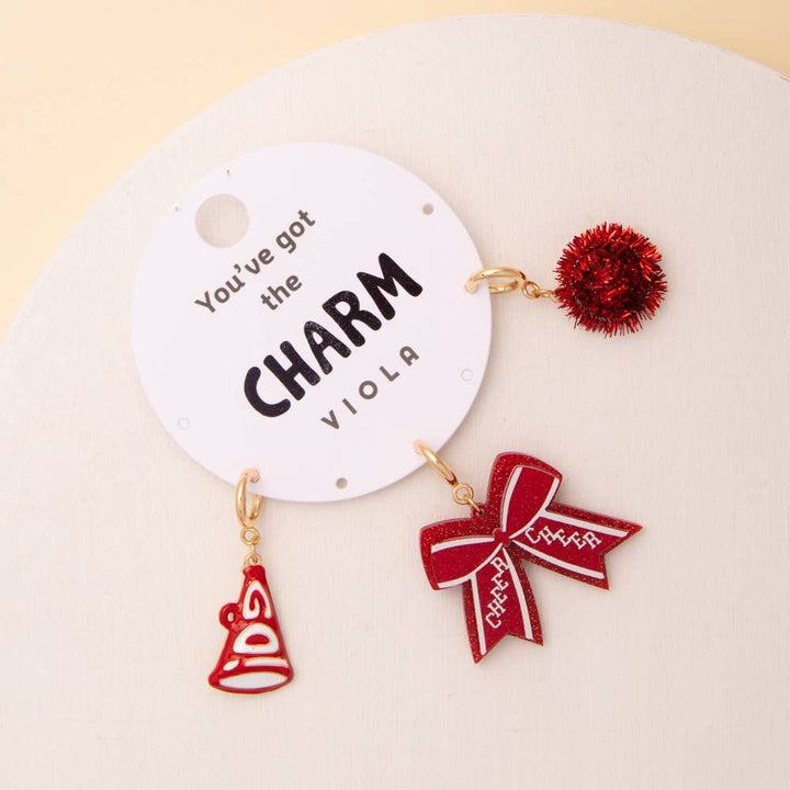 Red and gold charm set with pom pom, megaphone, glittered bow on card. Sizes: one size fits most.
