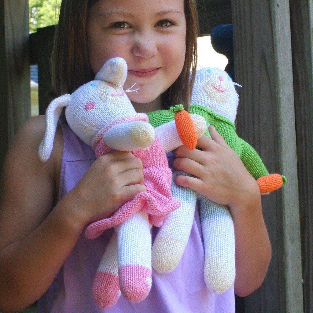 A young girl holding two Harriett the Bunny Knit Doll: 12" Plush dolls, one pink and one green, smiles warmly