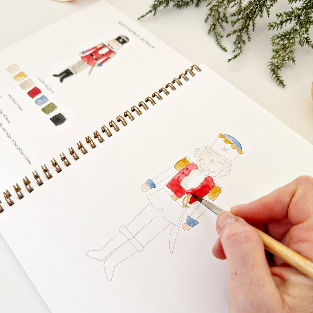 Red watercolor paint being applied to a nutcracker illustration in a Nutcracker watercolor workbook.