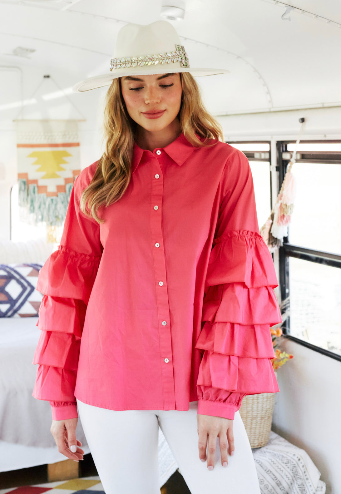 A woman in a Charlotte Button Down Ruffle Sleeve Top and white pants stands inside a sunny, bohemian-style camper van, her eyes gently closed under a stylish straw hat.