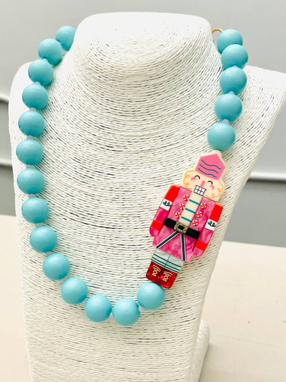 The HOLIDAY - Beaded Nutcracker Necklace (blue/pink) BNK1214 features large turquoise beads with a colorful Nutcracker pendant in pink, red, and blue at the center. Sizes available: please refer to product listing for options.