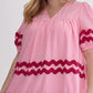 The Bay Ribbon Dress features ruffle sleeves and a wavy red rick rack trimming around the midsection. Sizes available are small, medium, large, and extra-large.