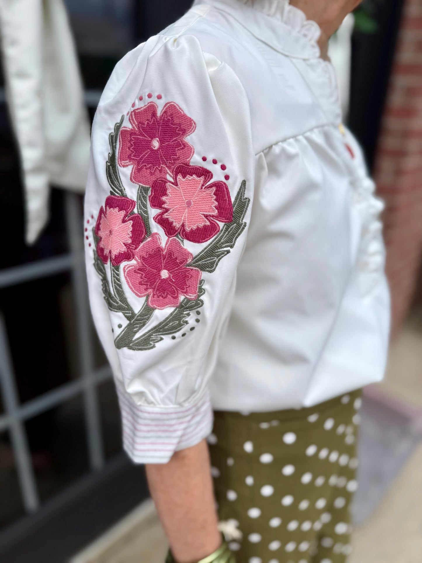 Emmie Ivory Top with ruffle neckline and pink floral embroidery on the sleeve. Available in sizes XS–XL.