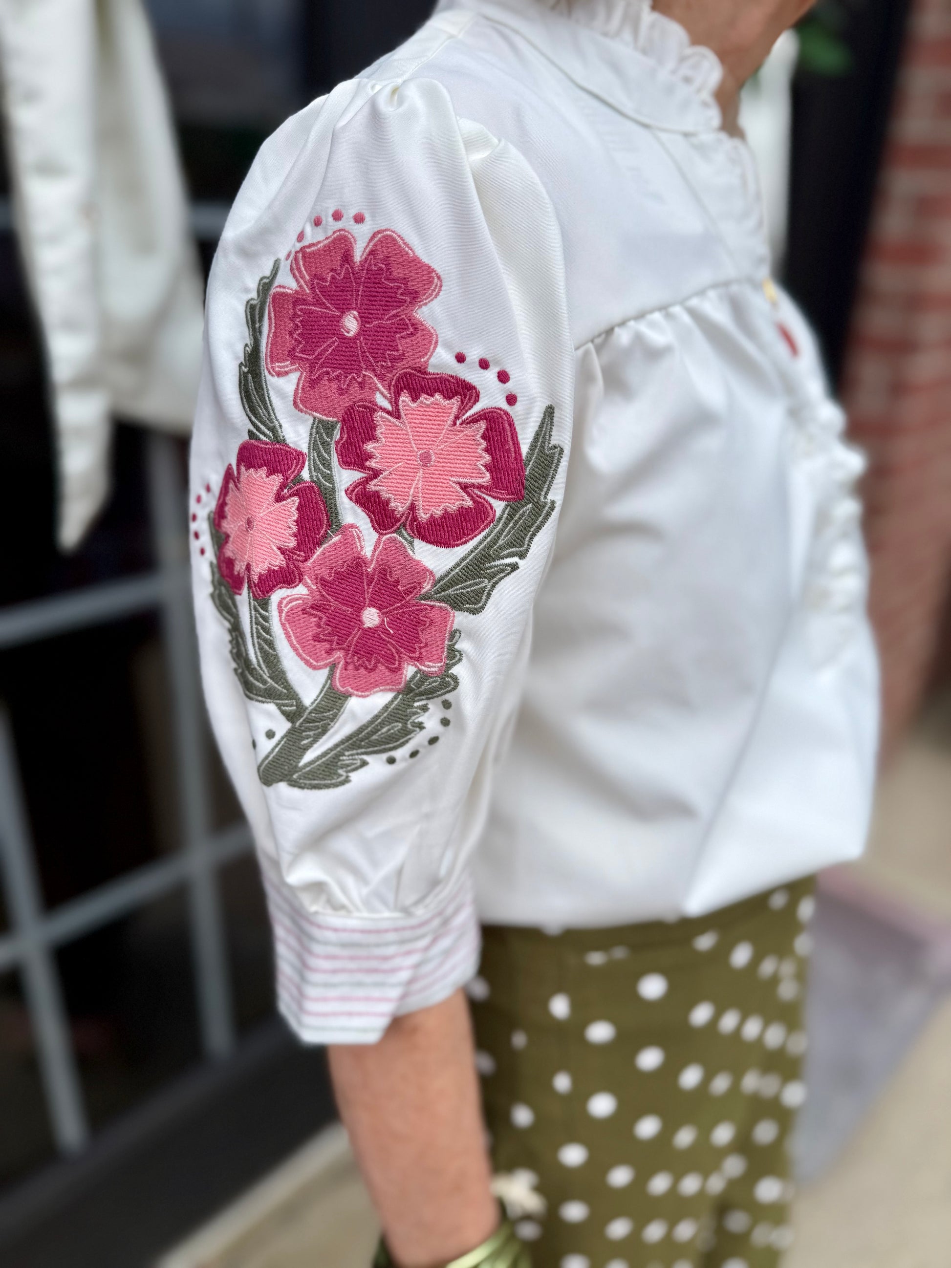 Emmie Ivory Top with ruffle neckline and pink floral embroidery on the sleeve. Available in sizes XS–XL.