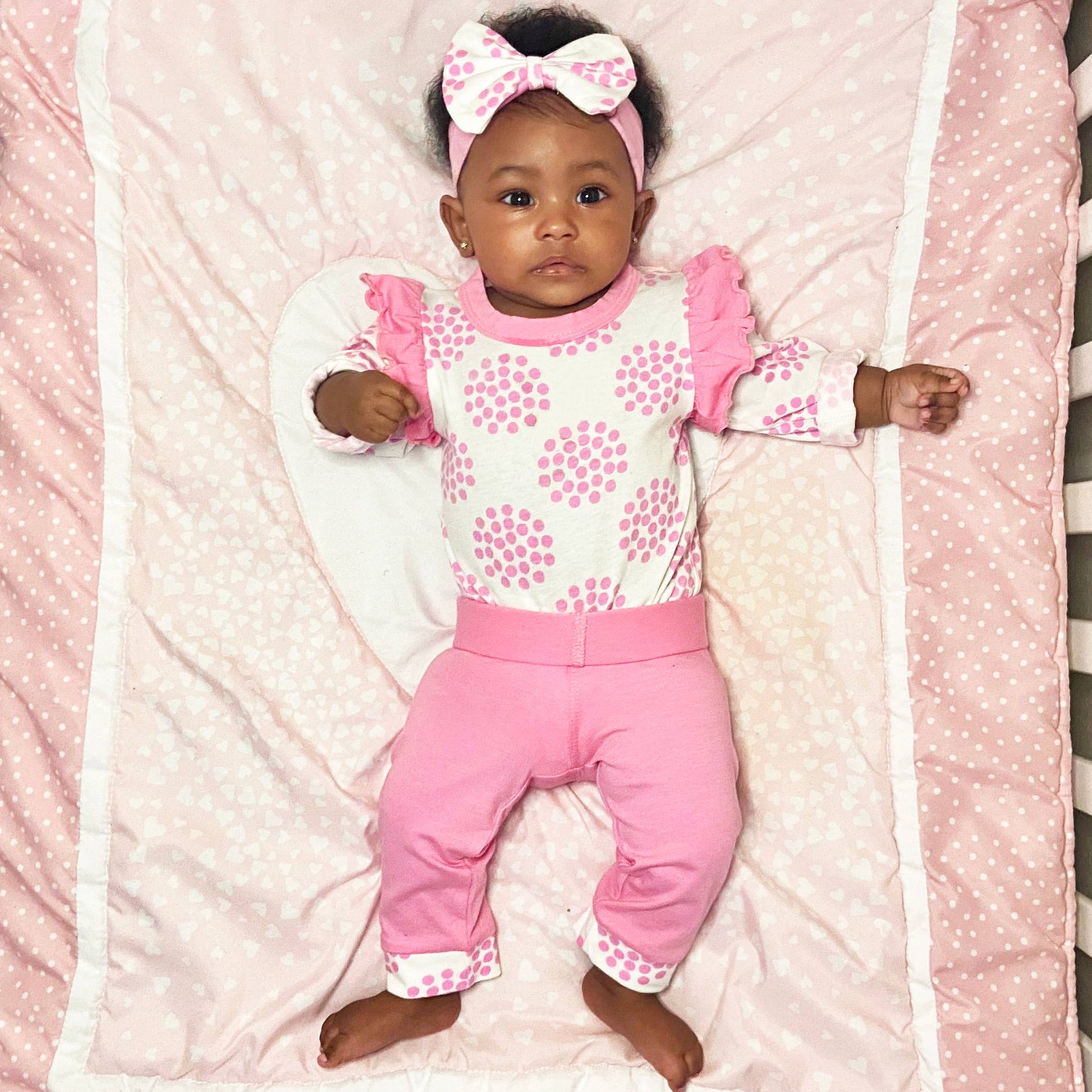 A baby with a bow in her hair is lying on a pink, polka-dotted blanket. She wears Layette Girls Baby clothes Shower Gift Pink Polka Dot Easter: 3-6 Mo top with ruffle sleeves and matching pants, looking up with