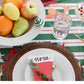 Festive holiday table setting with a Baby Bowl Pinstripes (available in small and medium sizes) filled with assorted fruits, a plate with a "Nora" name card, petite red stocking, glass of bubbly drink, water glass, salad, cookies, and cutlery on a green and white striped tablecloth.