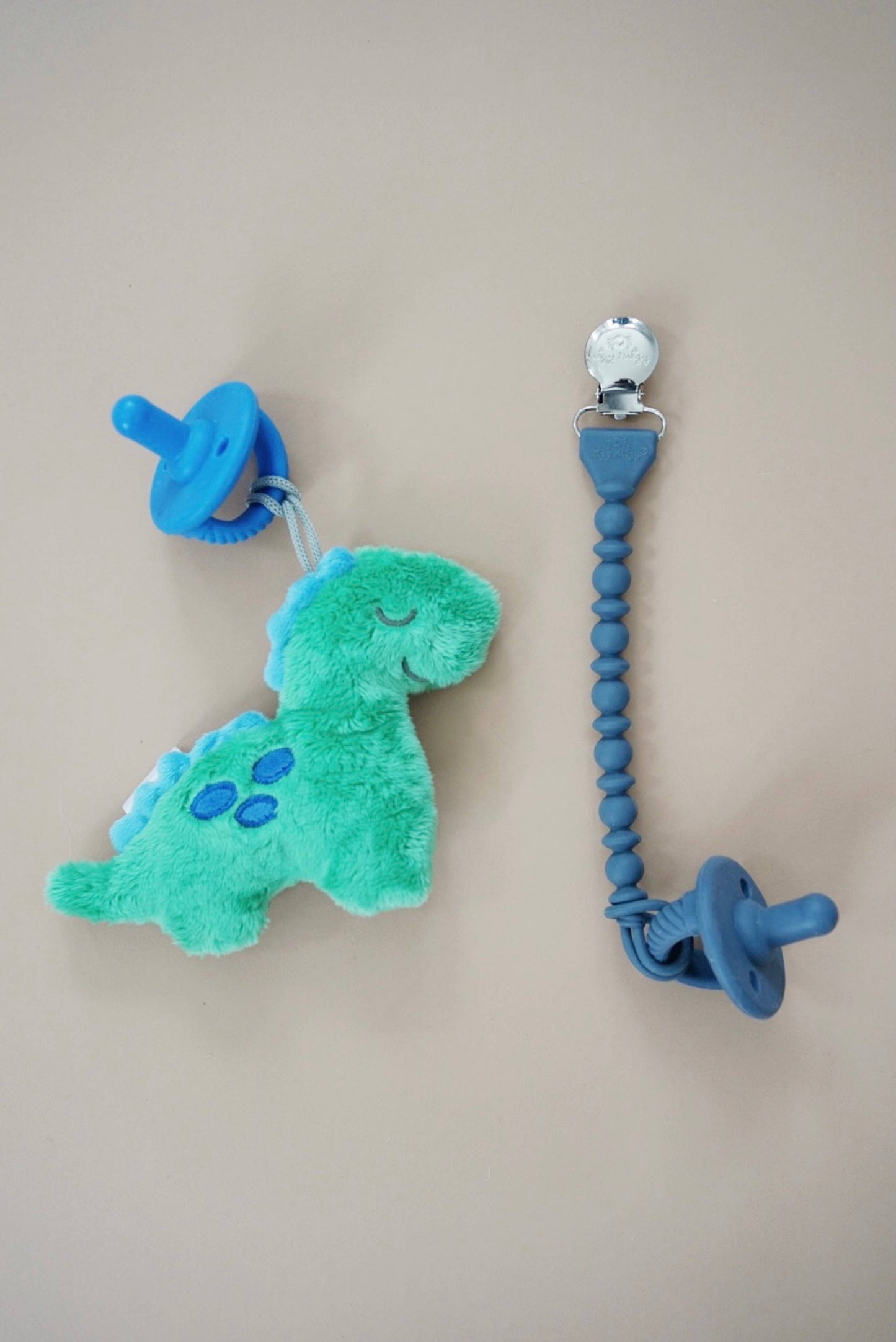 Two Sweetie Soother clips with attached pacifiers against a beige background; one features a Sweetie Pal™ Plush & Pacifier: Dino and the other, a blue silicone bead string.