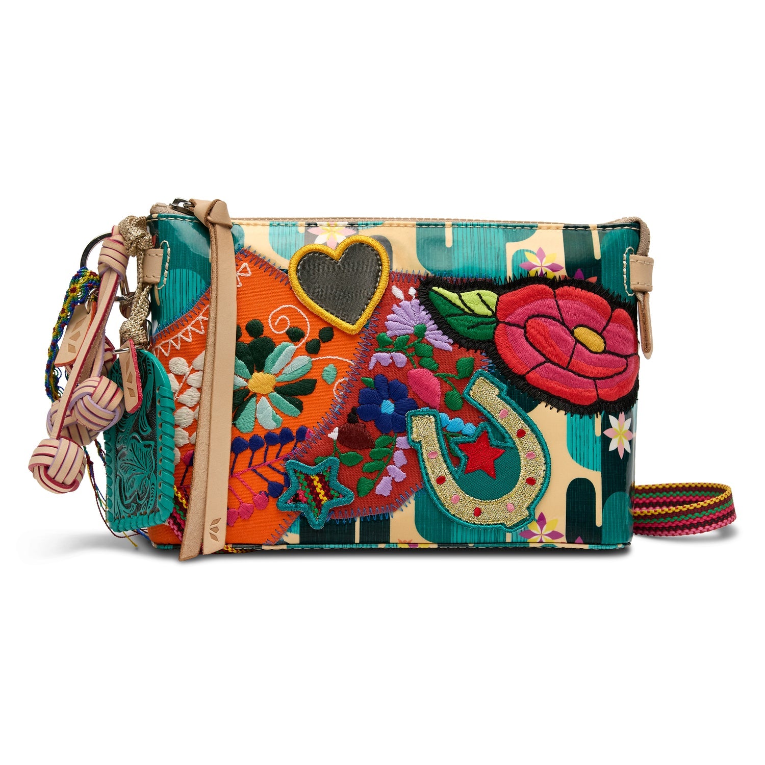 The Midtown Crossbody, Dezi is a colorful ConsuelaCloth bag with embroidered flower, horseshoe, heart, and star patches, a multicolored strap, and decorative tassels. Available in one size.