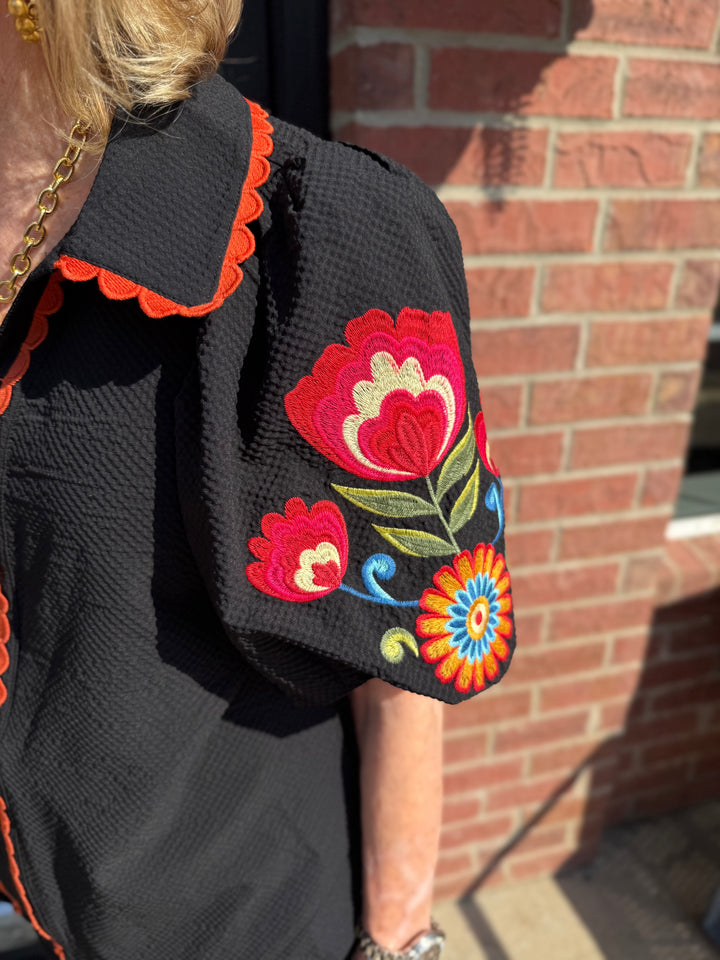 Black Fall in Bloom Blouse with orange trim and colorful floral embroidery on puff sleeves; available in sizes XS–XXL.