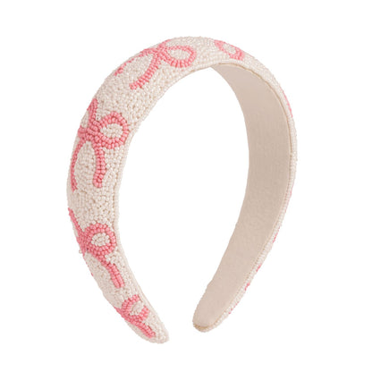 White fabric headband with small white beads, pink beaded swirls, and a hand-beaded bow. Available in one size.