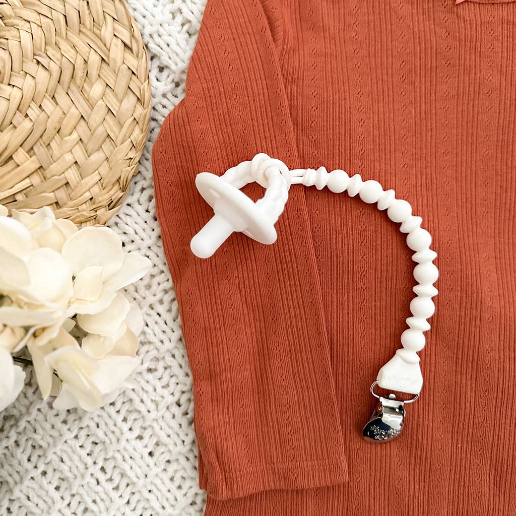 A Sweetie Strap™ Silicone One-Piece Pacifier Clip: Hero Blue Beaded with a white bead chain and airplane-shaped clasp lies on an orange textured cloth, beside a wicker object and white flowers on a knit surface.