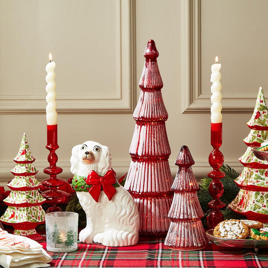 The holiday table display features red and green Christmas tree decorations, a porcelain dog figurine with a red bow, and holiday cookies. It includes Hand-Blown Glass Candlesticks for tall red candles and is completed by a red and plaid tablecloth. Available sizes: N/A.