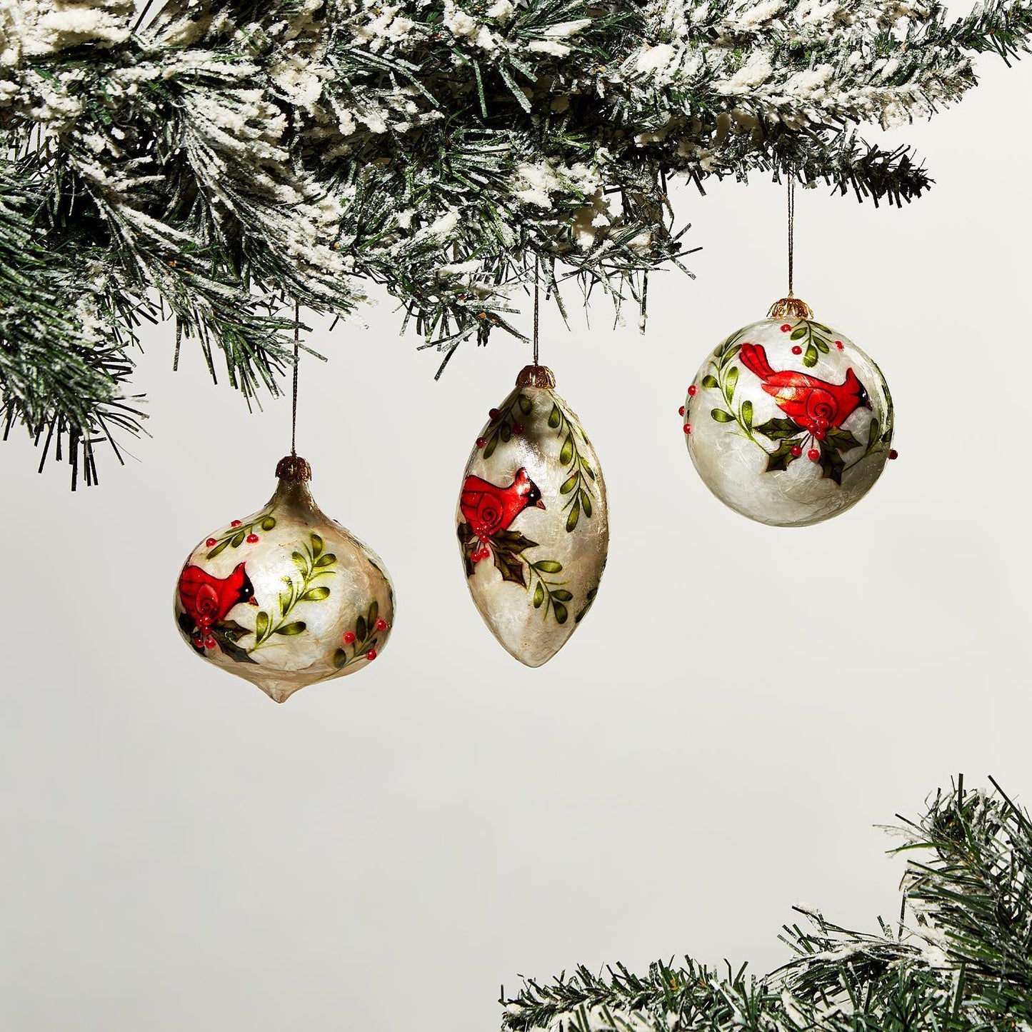 Three Hand-Painted Capiz Shell Cardinal Ornaments feature red bird and green foliage designs. Perfect for holiday décor, each ornament is meticulously painted. Sizes available: One size only.