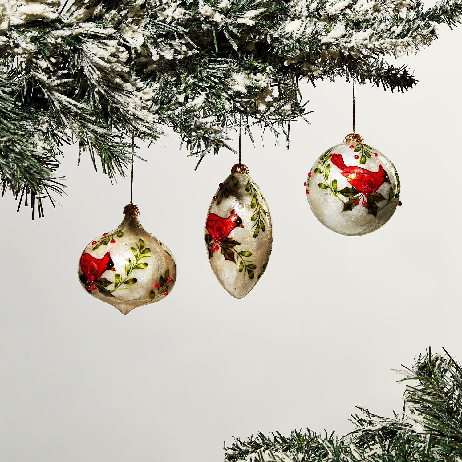 Three Hand-Painted Capiz Shell Cardinal Ornaments feature red bird and green foliage designs. Perfect for holiday décor, each ornament is meticulously painted. Sizes available: One size only.