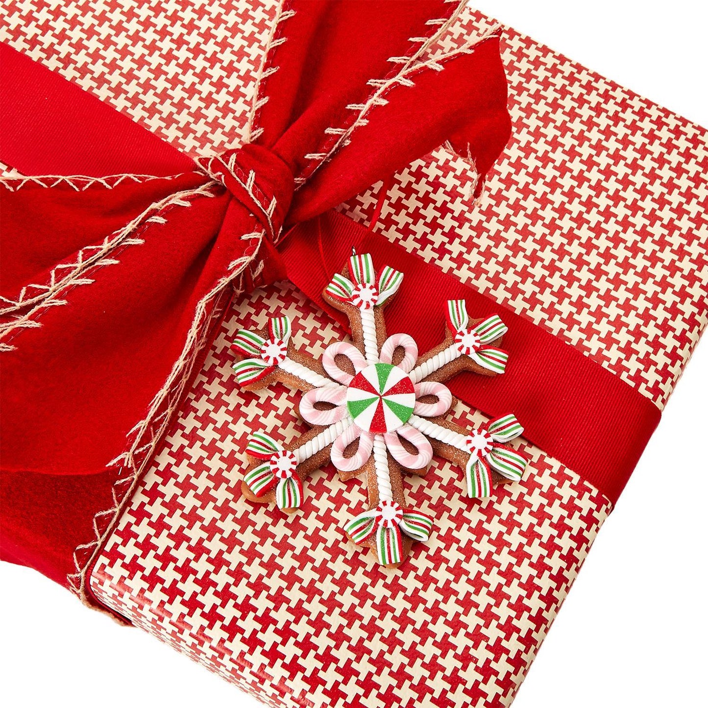 A present wrapped in red and white houndstooth paper with a red ribbon, topped with a Gingerbread Hand-Crafted Ornament featuring red, white, and green striped snowflake designs. Available sizes: Small, Medium, Large.