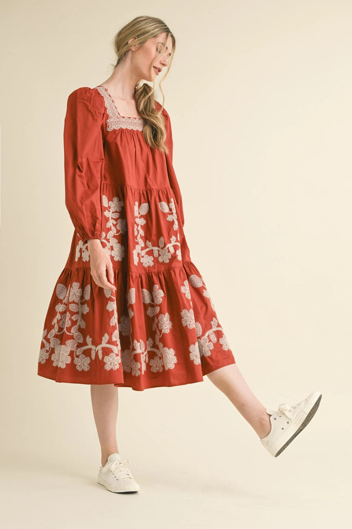 Rust midi dress with long sleeves, square neckline, and white floral embroidery on bodice and tiered skirt. Available in various sizes.