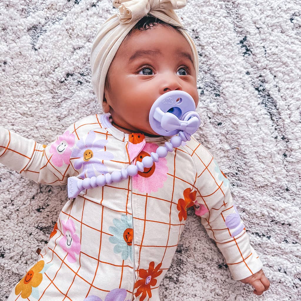 A baby wearing a floral printed onesie and a headband lies on a textured rug, looking upward with a purple pacifier attached by a Sweetie Strap™ Silicone One-Piece Pacifier Clip: Mint Braid in their mouth.