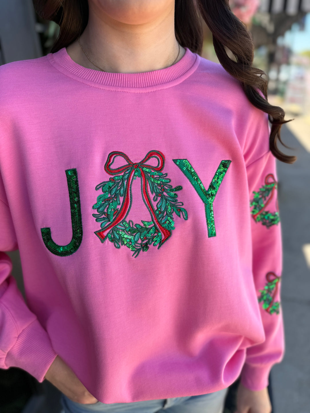 Pink Sarah Joy Wreath Sweatshirt with green sequined “JOY” lettering, a sequined wreath ‘O’, and a red bow. Available in all sizes.