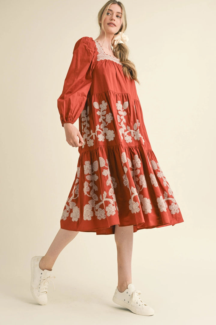 Rust midi dress with long sleeves, square neckline, and cream boho floral embroidery. Available in sizes XS-XL.
