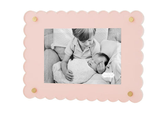 A black and white photo of a young boy gently touching a baby's head, both lying on a sofa, framed in a Scalloped Acrylic Frame with gold accents.