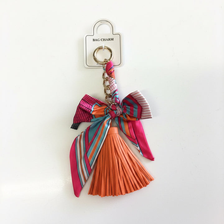 Gold keyring with pink and blue bow ribbon and orange leather tassel. Available in one size.