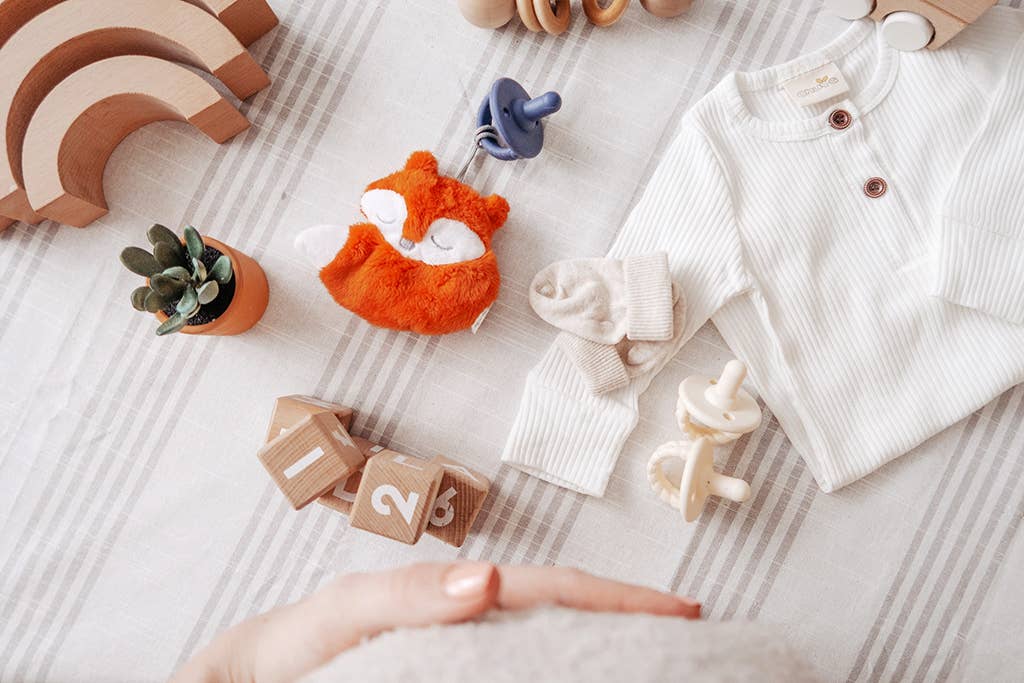 A cozy baby's nursery setup featuring a soft striped onesie, a sleeping fox plush toy, wooden toys, Sweetie Pal™ Plush & Pacifier: Dino, and a small potted plant, set against a textured wall.