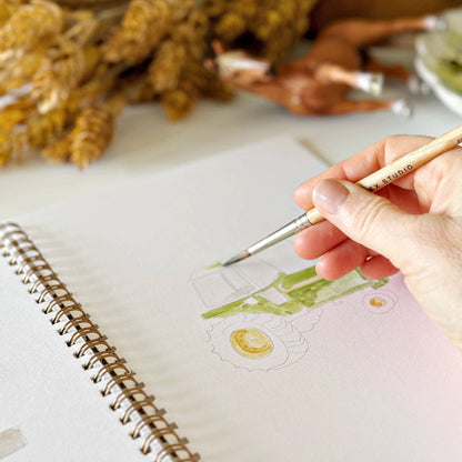 Hand painting a green tractor in the Farm Watercolor Workbook, spiral-bound coloring book. Available in multiple sizes.