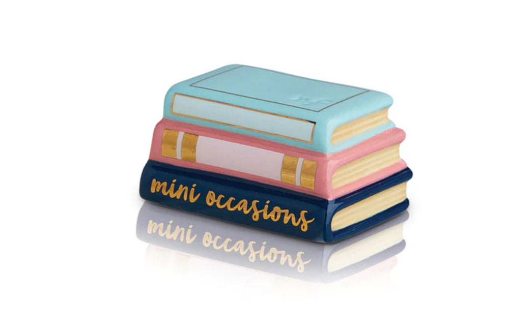 A stack of three ceramic books titled "Book and Mini Book w/Mini" showcasing "mini occasions" in gold lettering on the spine of the bottom book. Designed by Nora Fleming, these pastel-colored books feature light blue, peach, and navy hues. Available sizes: N/A (decorative set only).