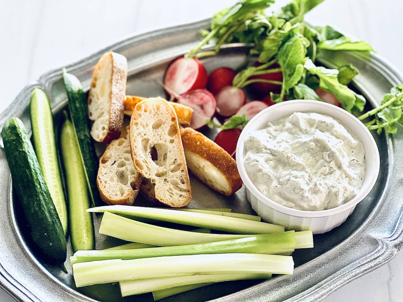 A healthy platter comprised of fresh, sliced cucumbers, celery sticks, radishes, and arugula. Accompanied by bread slices for dipping in a bowl of rich Artichoke Parmesan Dip Mix. All items are organized neatly on a portable metal tray. Available in small, medium and large sizes.