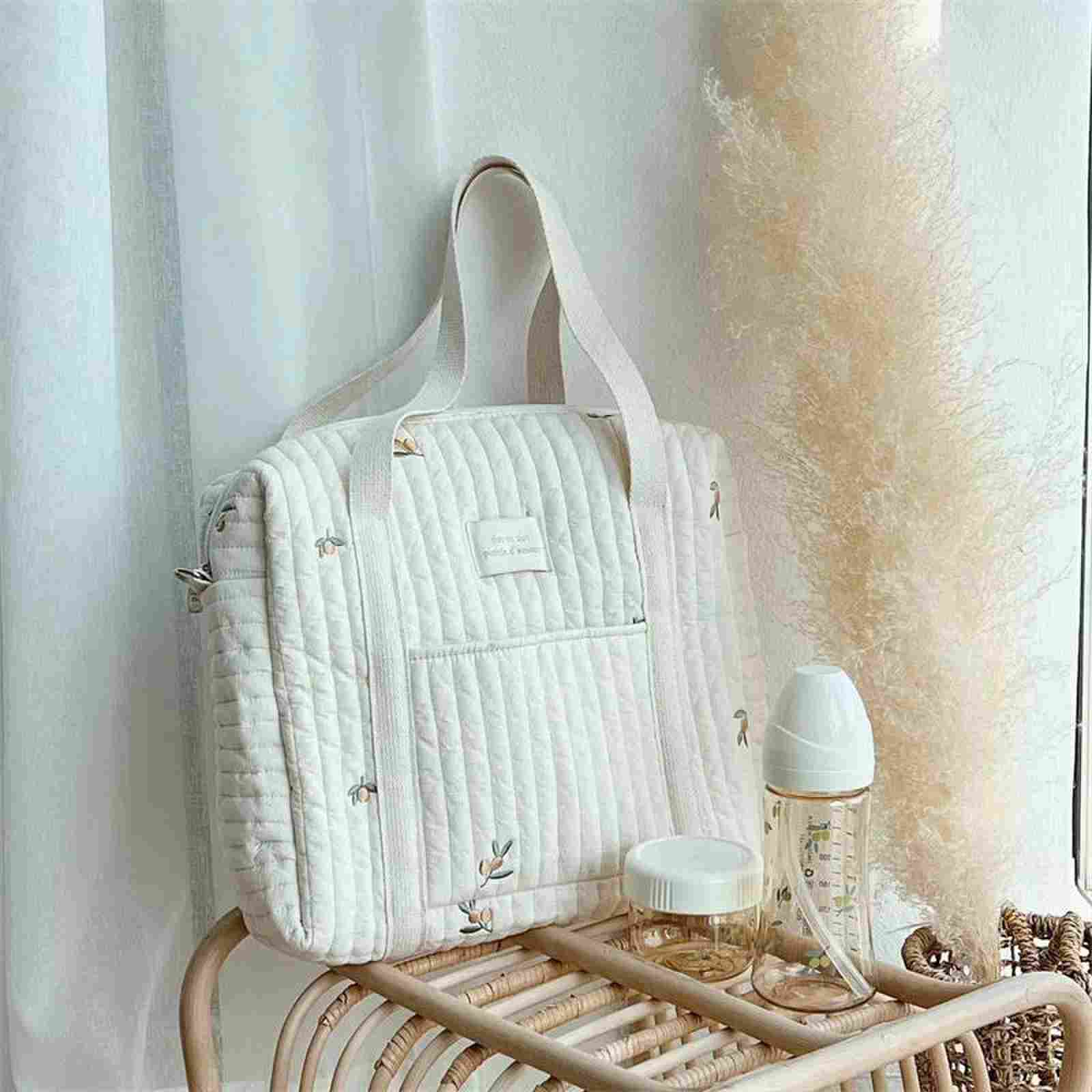 A Olive Embroidered Stroller and Diaper Bag in a soft beige color, displayed on a wooden rack next to a baby bottle and a small container, with stylish patterns and a neutral-toned, textured wall.