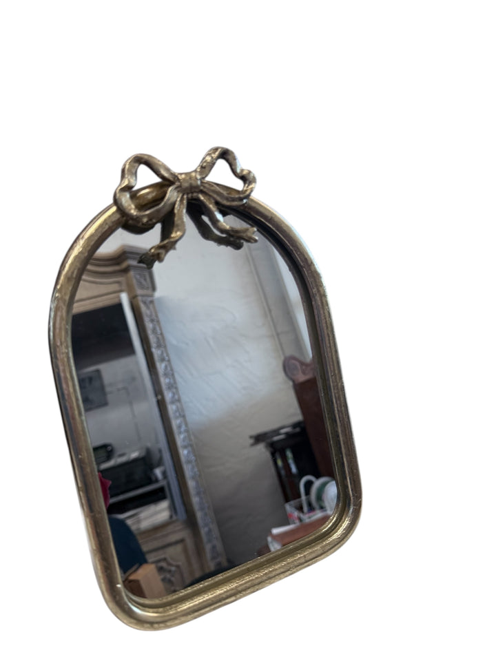 Small arched table mirror with a brass-tone frame and decorative bow on top. Available sizes not specified.