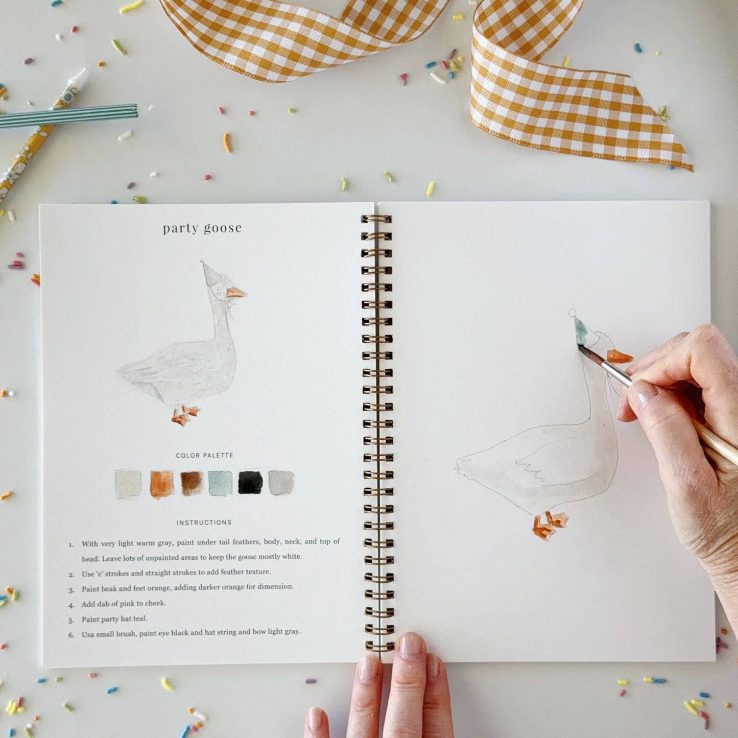 An open Celebrate watercolor workbook featuring a goose illustration, color palette, and beginner-friendly painting instructions. Available in multiple sizes.