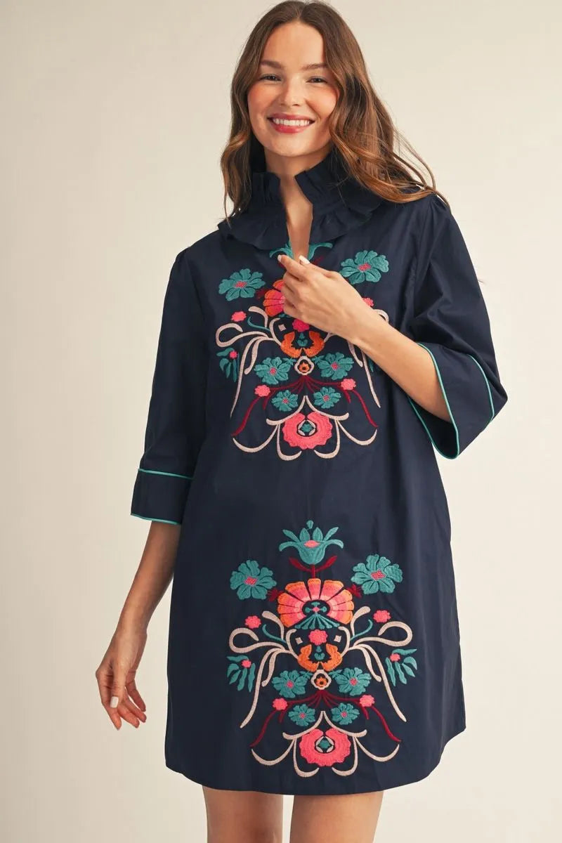 Navy mini dress with colorful floral embroidery, three-quarter sleeves with light blue piping, and a delicate ruffled neckline. Available in sizes XS–XL.