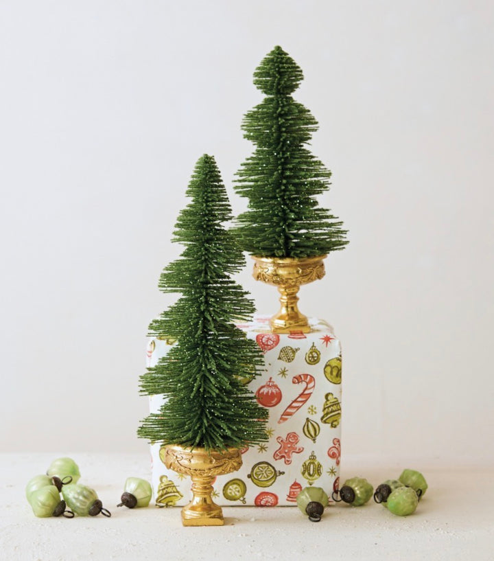 Two green glittered bottle brush trees on gold pedestal stands.
